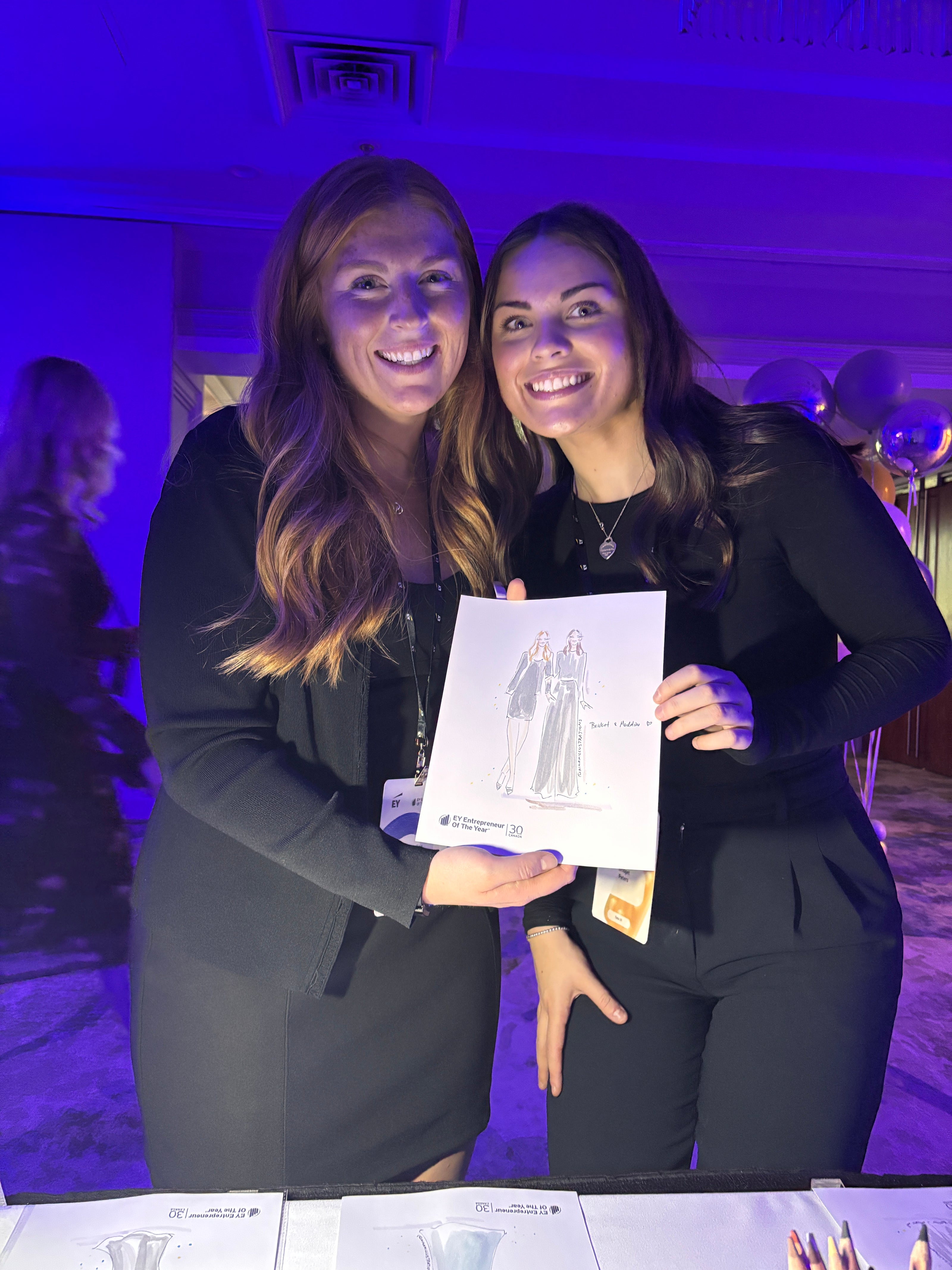Two women holding Nina Maric illustration in a room with purple lighting
