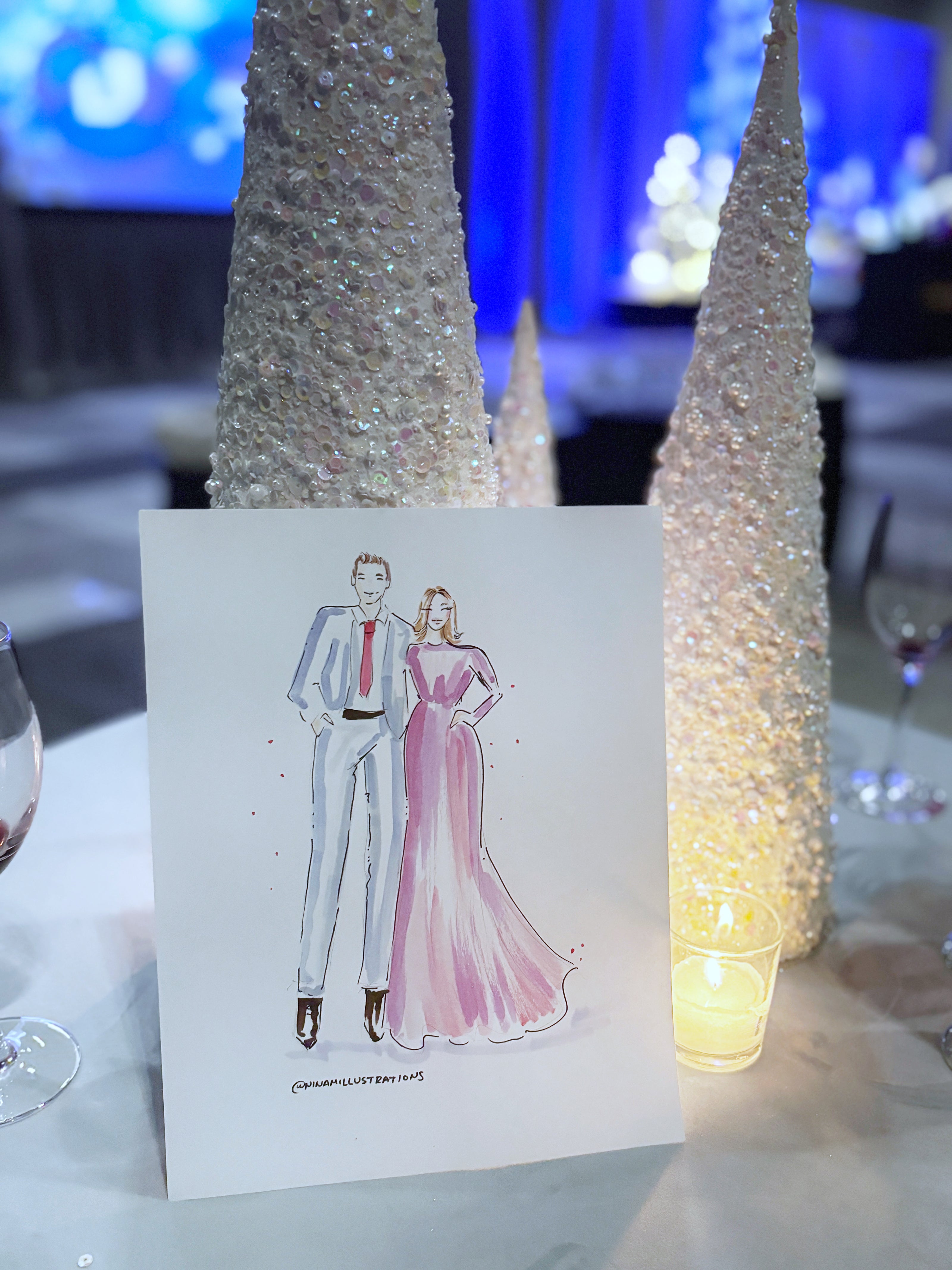Card with a drawing of a couple on a table with decorative trees and lights.