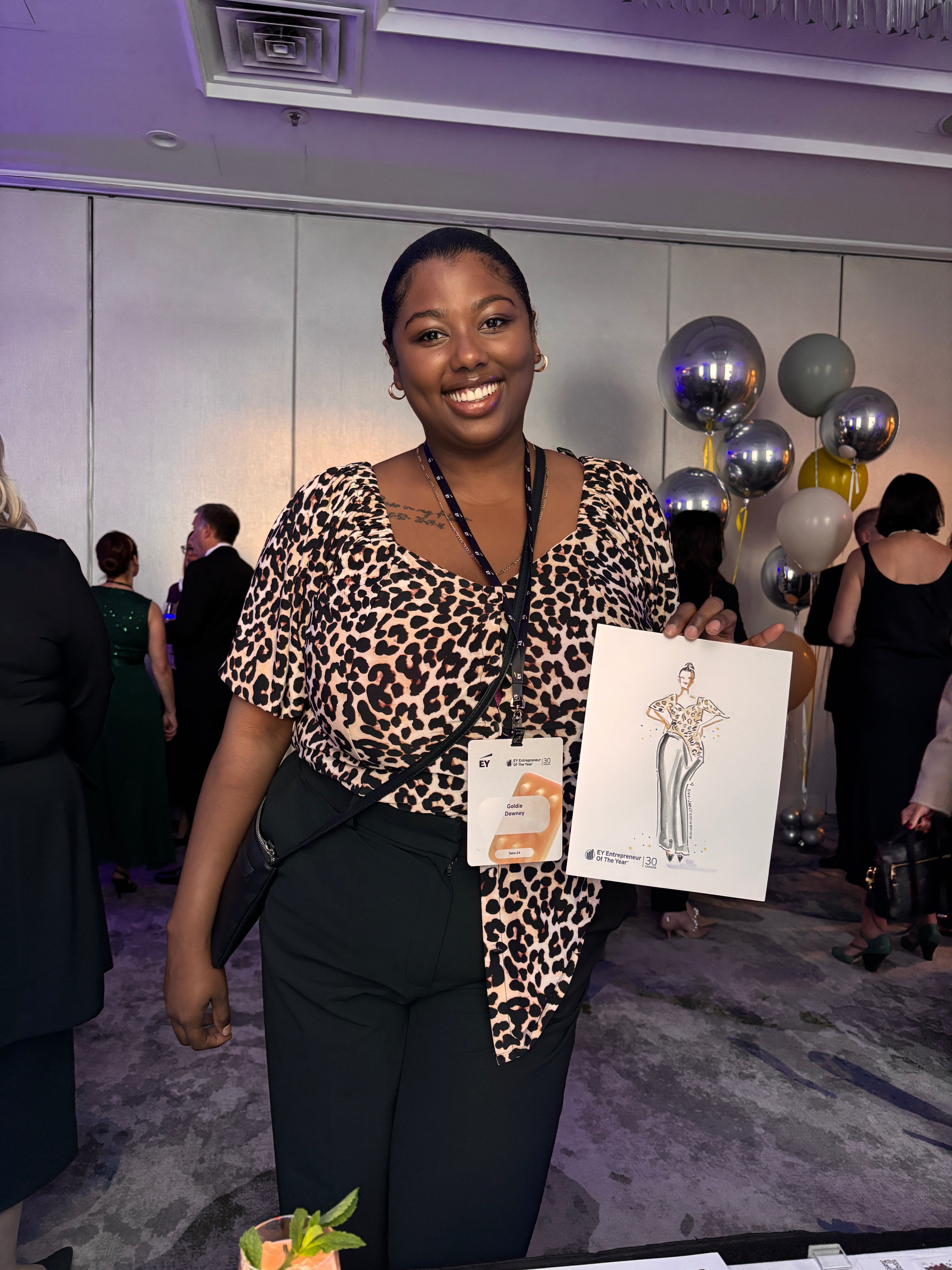 Woman in leopard print top holding a Nina Maric drawing at an event with balloons and people in the background.