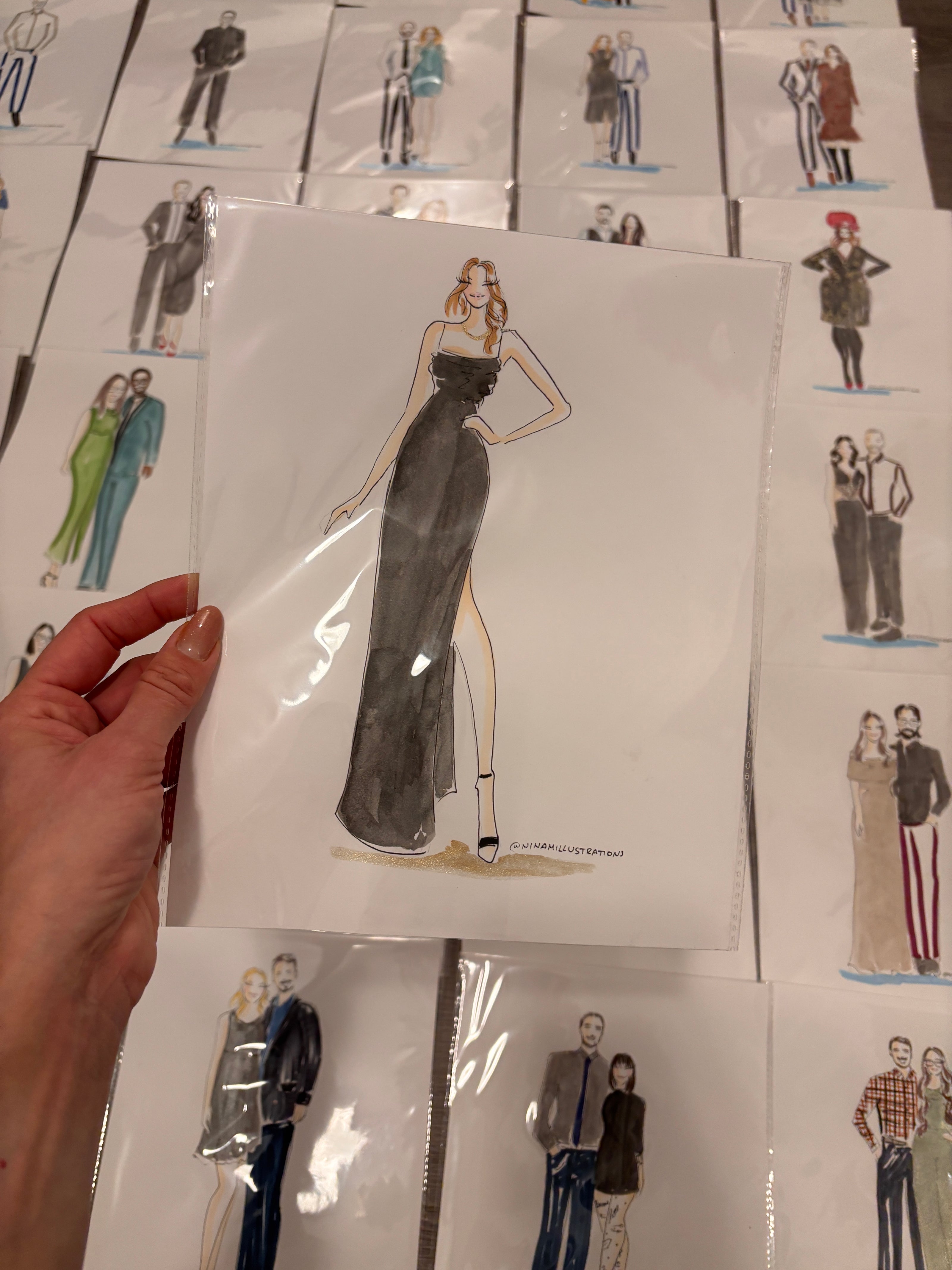 Hand holding a piece of paper with a drawing of a person in a dress, surrounded by other similar drawings on a white surface.