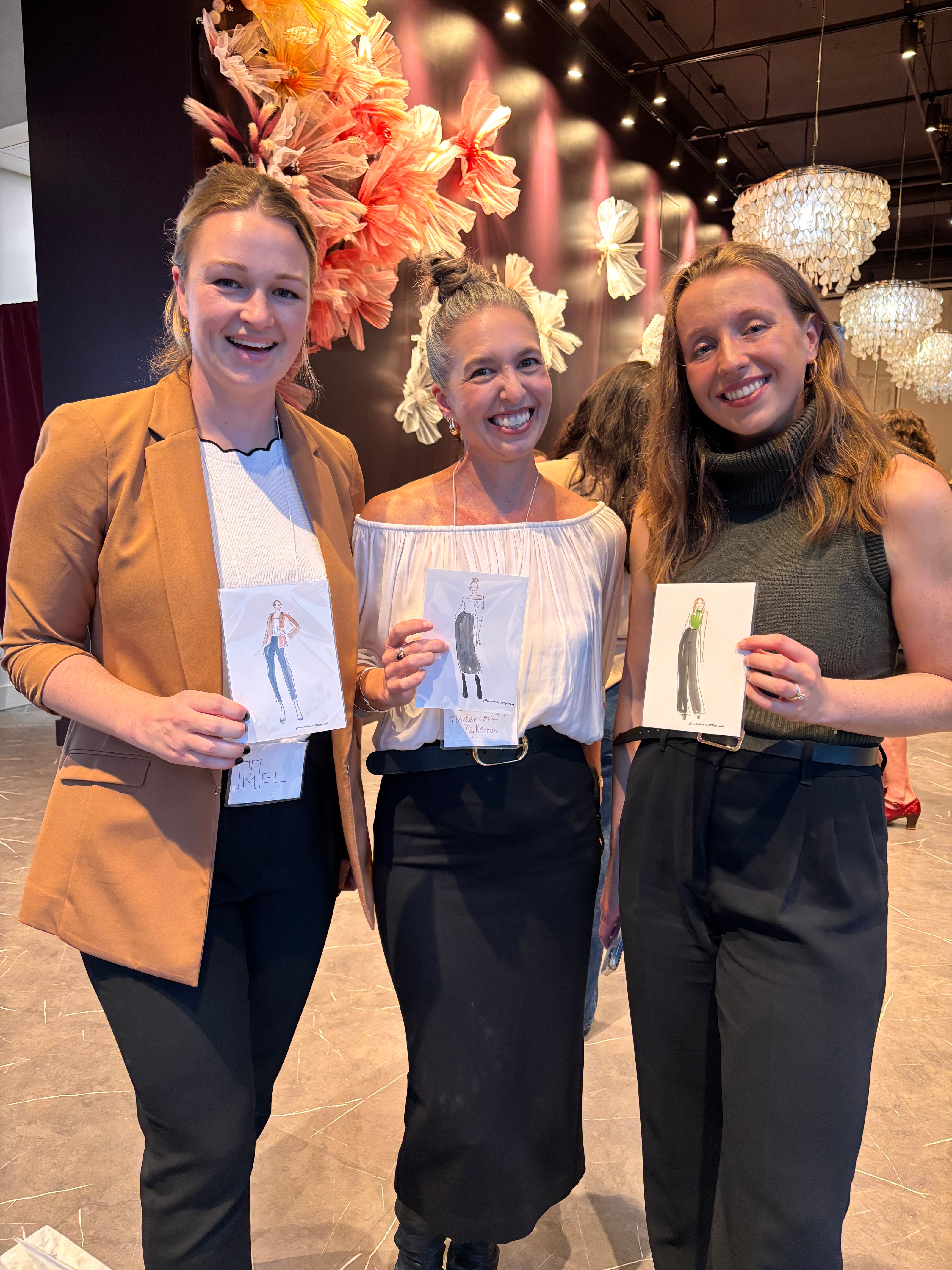 Three women standing together, each holding a card with Nina Maric illustrations on it, in an indoor setting.