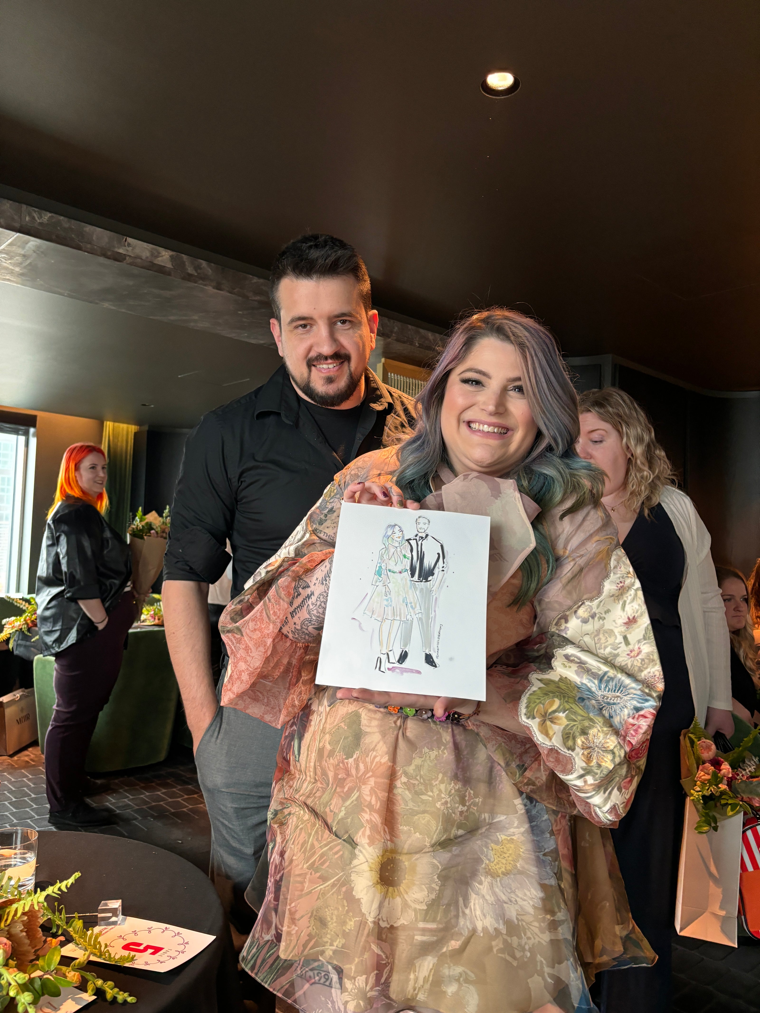 Two people standing next to a table with a Nina Maric drawing on it, in a casual indoor setting.