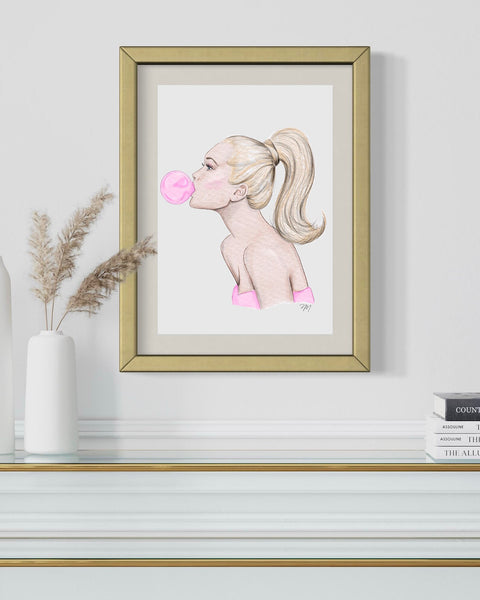 Nina Maric art in a gold frame on display, featuring woman blowing pink bubble